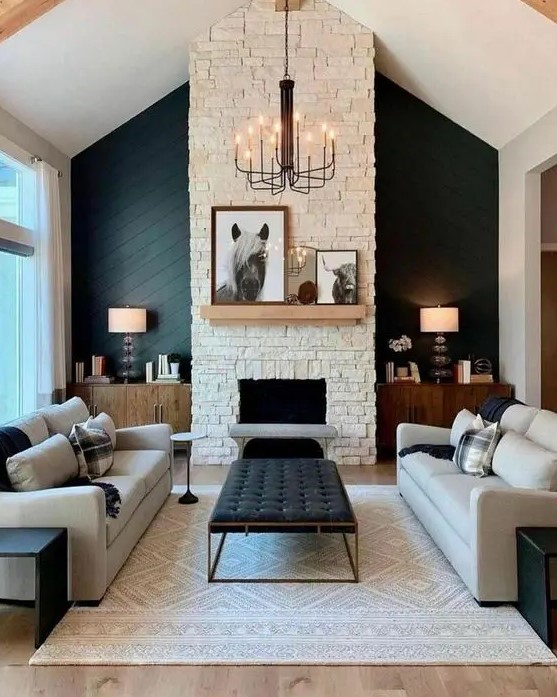 a sophisticated modern barn living room with a fireplace clad with faux stone, neutral seating furniture, a printed rug, a black ottoman, table lamps and a chic chandelier
