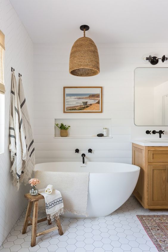 a stylish modern farmhouse bathroom done in neutrals, with shiplap on the walls, white hex tiles, a timber vanity, a mirror, a woven pendant lamp and an oval tub