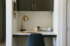 a tiny home office built into a small nook, with a built-in desk and a large cabinet, a navy chair, a brass table lamp
