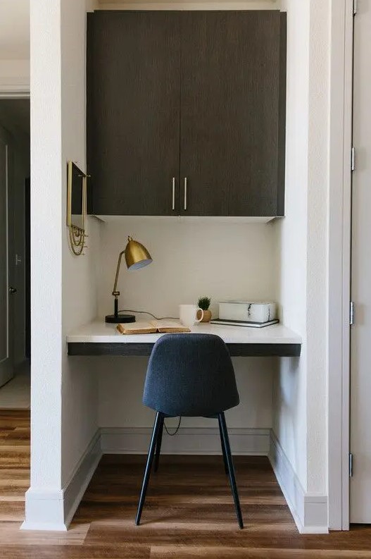 A tiny home office built into a small nook, with a built in desk and a large cabinet, a navy chair, a brass table lamp