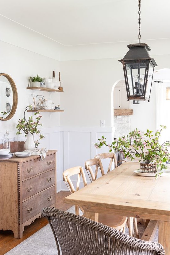 A welcoming farmhouse dining room with a light stained table and chairs, wicker chairs, a stained credenza, metal lanterns and greenery
