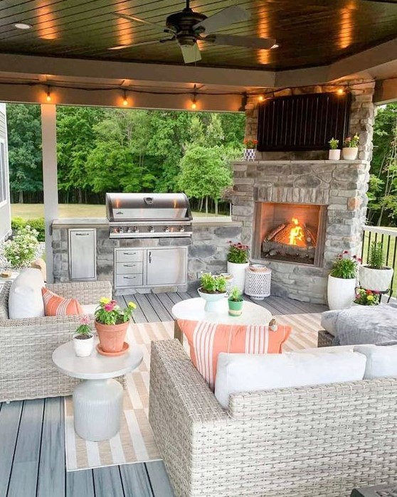 a welcoming outdoor living room done in modern farmhouse style, with a fireplace, a grill, neutral wicker furniture and printed textiles