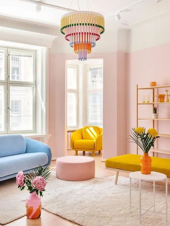 a welcoming pastel living room with pink walls, a blue sofa, a yellow chair, a yellow daybed, a pink pouf and a colorful chandelier