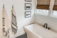 a white modern farmhouse bathroom with shiplap walls, a tub, windows with shades, a star-shaped pendant lamp and a penny tile floor