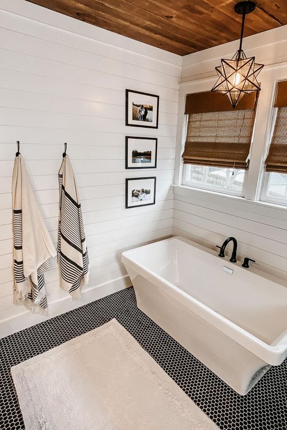 A white modern farmhouse bathroom with shiplap walls, a tub, windows with shades, a star shaped pendant lamp and a penny tile floor