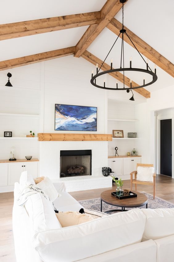A white modern farmhouse living room with wooden beams, a fireplace, built in shelves and cabinets, a creamy sofa and a black chandelier