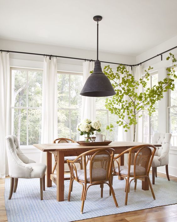 an airy dining room with a stained table and rattan chairs, a black pendant lamp, a potted tree and a neutral rug