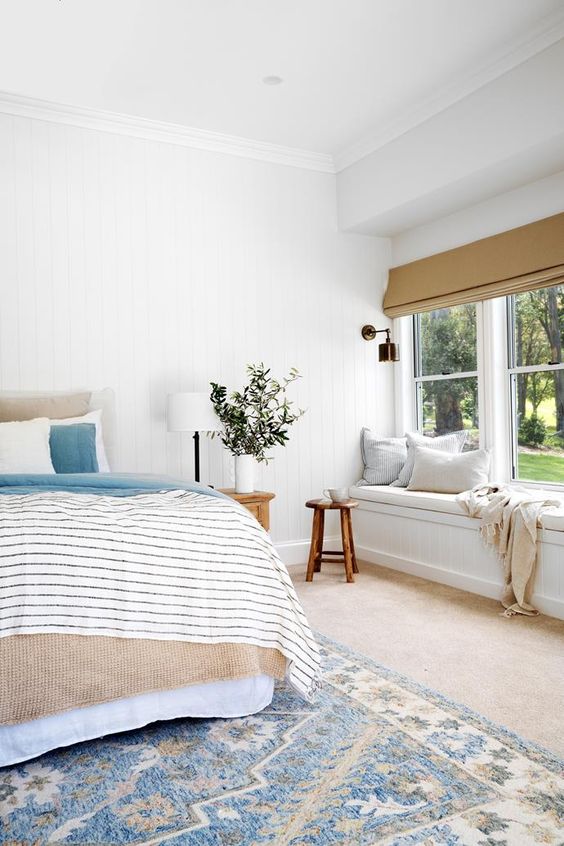 an airy modern farmhouse bedroom with a windowill daybed, a bed with neutral and printed bedding, wooden stools and greenery