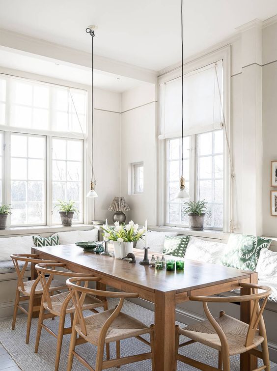 An airy modern farmhouse dining room with a large built in bench, a stained table and chairs, green and white pillows and pendant lamps