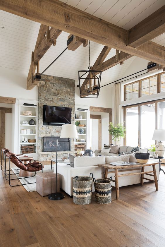 an airy modern farmhouse living room with wooden beams, a fireplace clad with stone, neutral seating furniture, leather chairs and a wooden bench