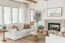 an airy modern farmhouse living room with wooden beams, a fireplace, white sofas and a wooden coffee table plus some baskets