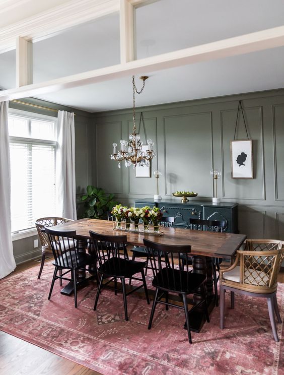 an elegant farmhouse dining room wiht green paneled walls, a dark green credenza, a stained table, black and stained chairs, a chic chandelier