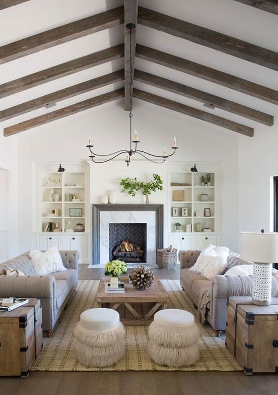 An exquisite barn living room with stained wooden beams, a brick fireplace and built in bookcases, grey sofas, a wooden coffee table and neutral poufs