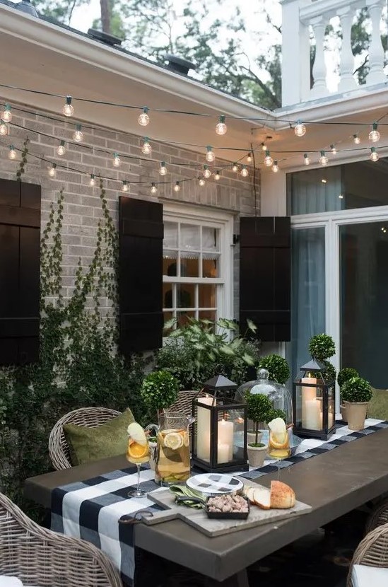 an inviting modern country patio with a table and wicker chairs, muted color pillows, potted greenery and candle lanterns plus lights over the space