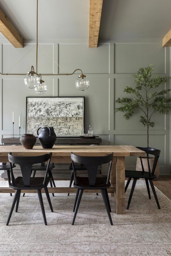 an olive green farmhouse dining room with paneled walls, a stained table, black chairs, a dark-stained sideboard and a chic chandelier