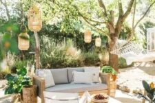 an outdoor living room with a grey sofa, neutral chairs, a low coffee table, lanterns hanging over the space, potted plants