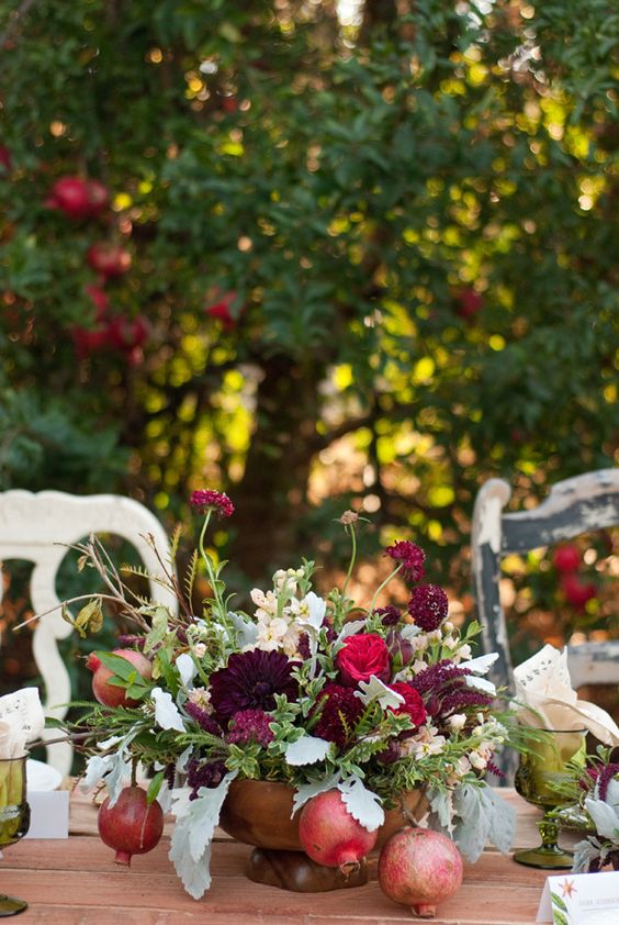 a bold fall or Thanksgiving centerpiece of red, burgundy blooms and greenery and pomegranates on the table is amazing