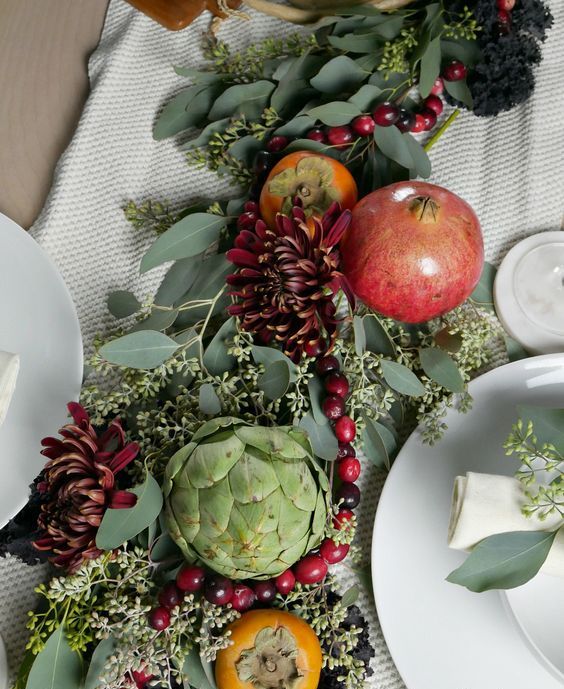 a fall or Thanksgiving table runner of greenery bold dahlias, an artichoke, fruit including pomegranates