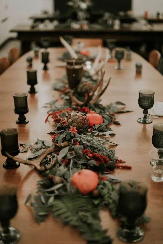 a fall table runner with greenery and leaves, pomegranates, antlers and artichokes plus candleholders is chic