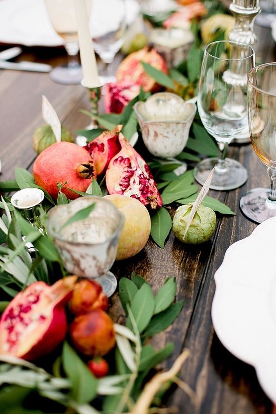a fall table runner with greenery, apples and pomegranates including cut ones, with candleholders