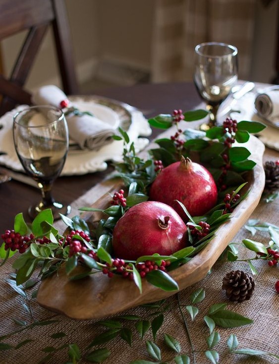 a rustic table with a burlap runner, greenery, berries, pinecones and a dough bowl with pomegranates and berries