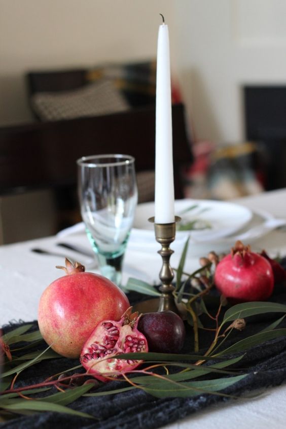 a simple and pretty fall centerpiece of a table runner, greenery, pomegranates and plums plus a tall candle