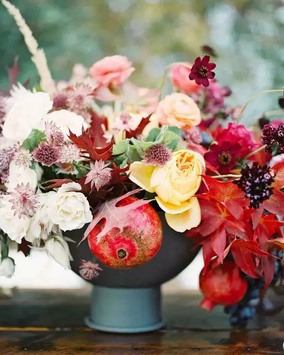 an adorable fall centerpiece of a grey vase, yellow, pink and deep red blooms, fall leaves and pomegranates that help to embrace the fall