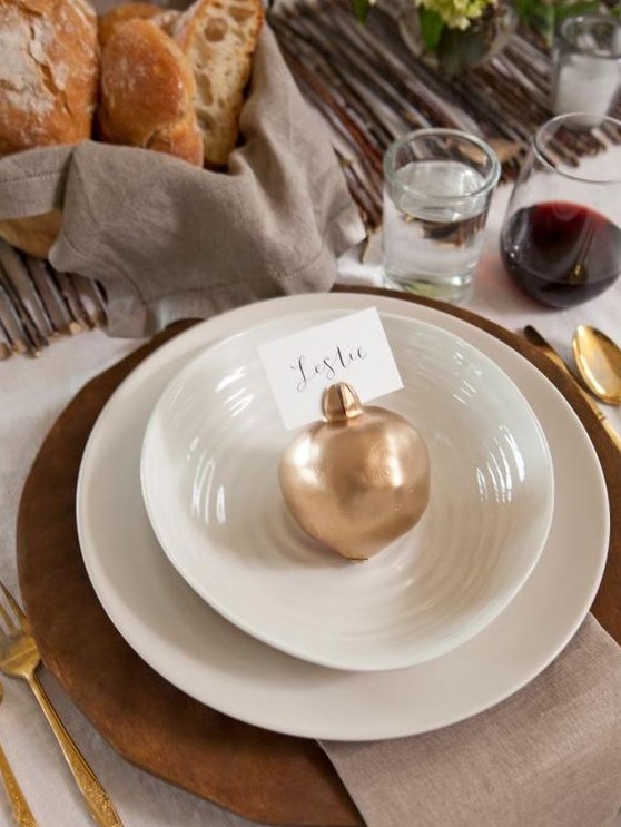 a chic fall place setting with a wooden placemat, white plates, a neutral napkin and a gilded pomegranate