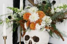 a Halloween decoration of a skull with white and orange blooms, black flowers, greenery and herbs is a nice centerpiece