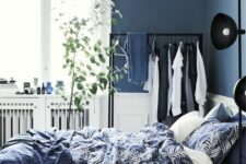 a Scandinavian bedroom with navy walls, a bed with navy and white bedding, a makeshift closet and a pendant lamp