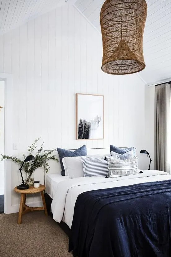 a beautiful and contrasting coastal bedroom with a white bed, navy and white bedding, a wooden stool and eucalyptus and a woven pendant lamp