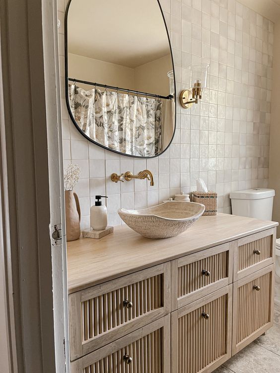 A beautiful neutral bathroom with Zellige tiles, a large reeded vanity, a bowl sink, an irregular shaped mirror and some chic decor