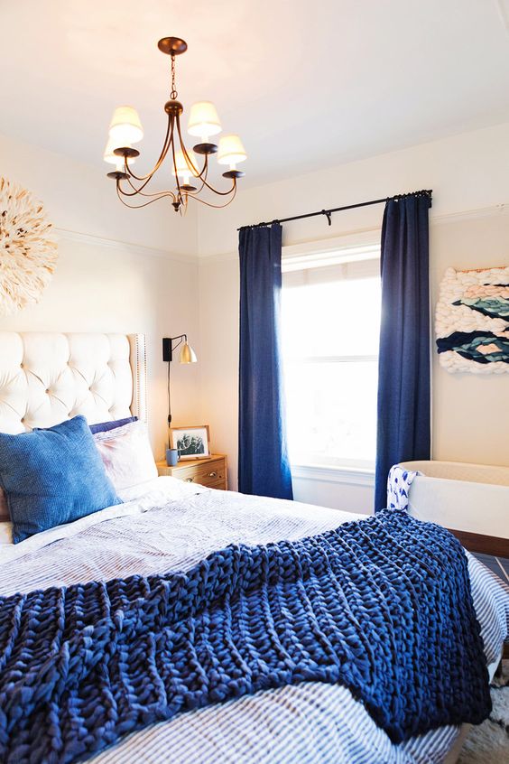a bold bedroom with a white bed, navy and white bedding, navy curtains, a macrame artwork and a crib