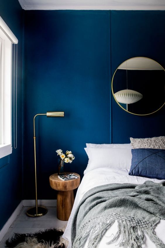 a bold navy bedroom with a bed and blue and white bedding, a round mirror, a wooden nightstand and a lamp