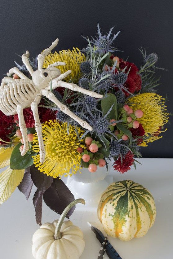 a bright floral arrangement with mustard, red blooms, berries and thistles plus a large spider on top