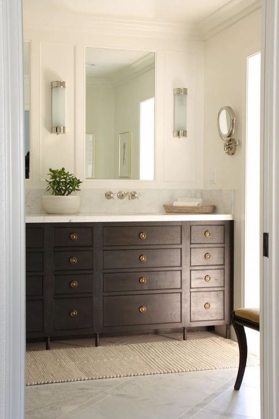 A chic and refined bathroom with a dark stained vanity, a mirror, some sconces and a vintage chair, a wall mirror