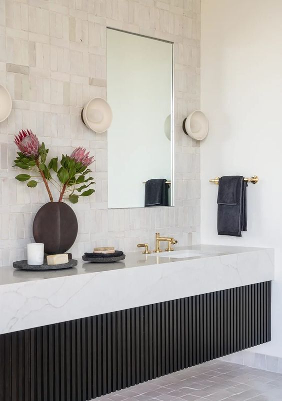 a chic bathroom with neutral tiles, a black fluted built-in vanity with a stone slab and gold fixtures