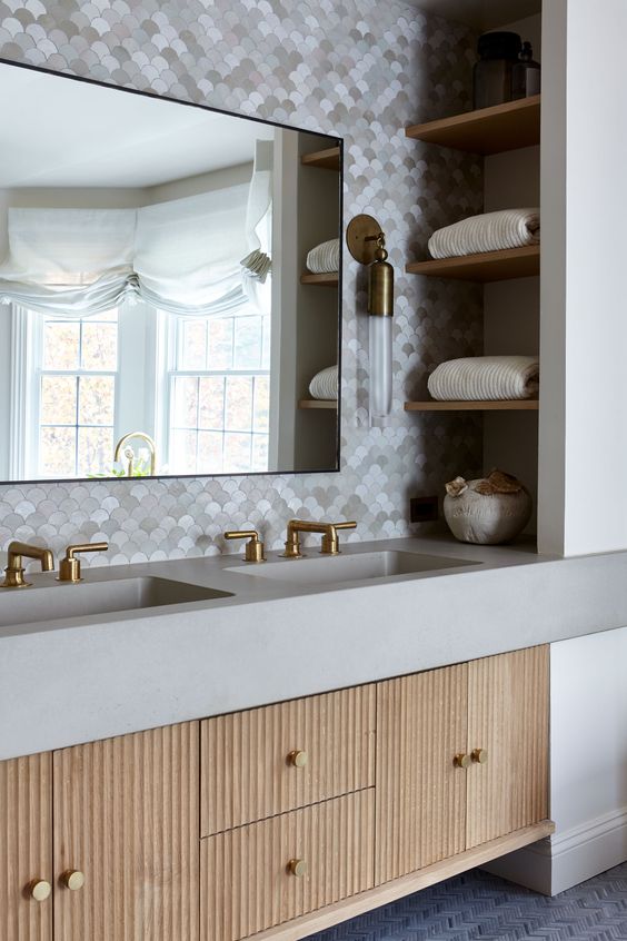 A chic bathroom with scallop tiles, a built in fluted vanity, niche shelves, a large mirror and elegant brass sconces