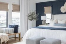 a chic bedroom with a navy paneled accent wall, a white bed and blue and white bedding, a woven bench with pillows