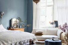 a chic bedroom with blue walls, a bed with neutral bedding, a dark green bench, a blue ottoman and a white sofa