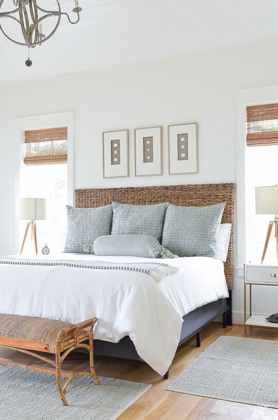 a chic coastal bedroom with a blue bed with a wicker headboard, a rattan bench, a wall gallery with seashells and a vintage chandelier