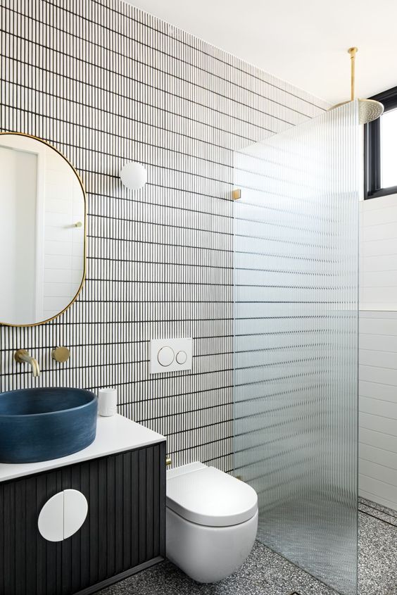 a chic modern bathroom with skinny tiles and grey terrazzo, a black fluted vanity, a navy vessel sink and an oval mirror