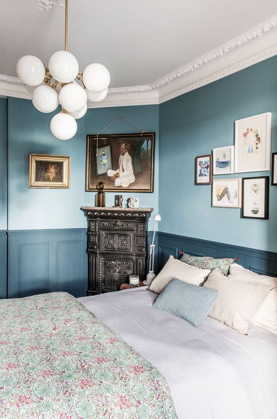 A chic vintage inspired bedroom with light blue walls, navy paneling, a ceiling with molding, a bubble chandelider and a vintage hearth