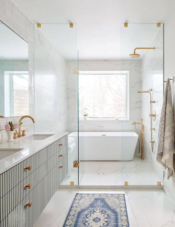 a chic white marble bathroom with a shower and a bathtub, a fluted vanity with two sinks, brass knobs and a printed rug