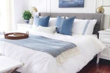 a coastal bedroom with a grey upholstered bed, blue and white printed textiles, a sea artwork, a stool and potted plants