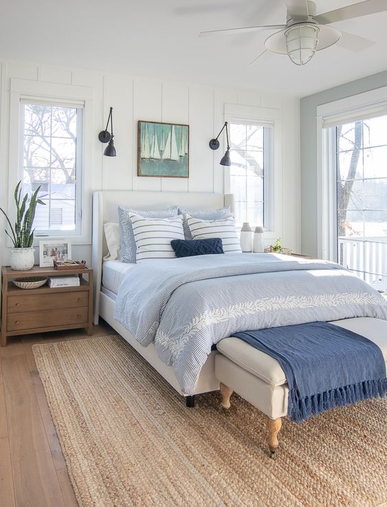a coastal bedroom with shiplap walls, a white bed with blue and white bedding, a white bench with a blue blanket and a seagrass rug