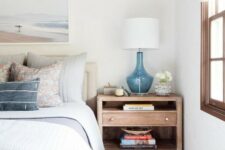 a coastal bedroom with white walls, a wooden floor and furniture, blue printed bedding, a sea artwork and a blue lamp