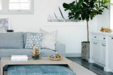 a coastal living room with a pale blue sofa, a wooden coffee table, a blue ottoman, a potted tree and printed pillows