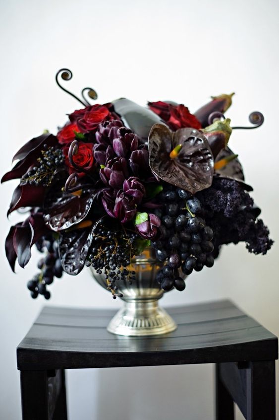 a dark and decadent Halloween wedding centerpiece of purple and black blooms, red roses, grapes and an eggplant