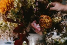 a dark and moody floral centerpiece of a silver urn, rust and mustard blooms, dried leaves and bright fall leaves